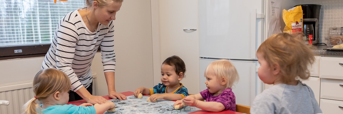 Neljä lasta pöydän ääressä leipomassa sämpylöitä aikuisen kanssa