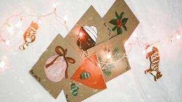 Itse tehtyjä joulukortteja pöydällä piparkakkujen ja jouluvalojen keskellä.