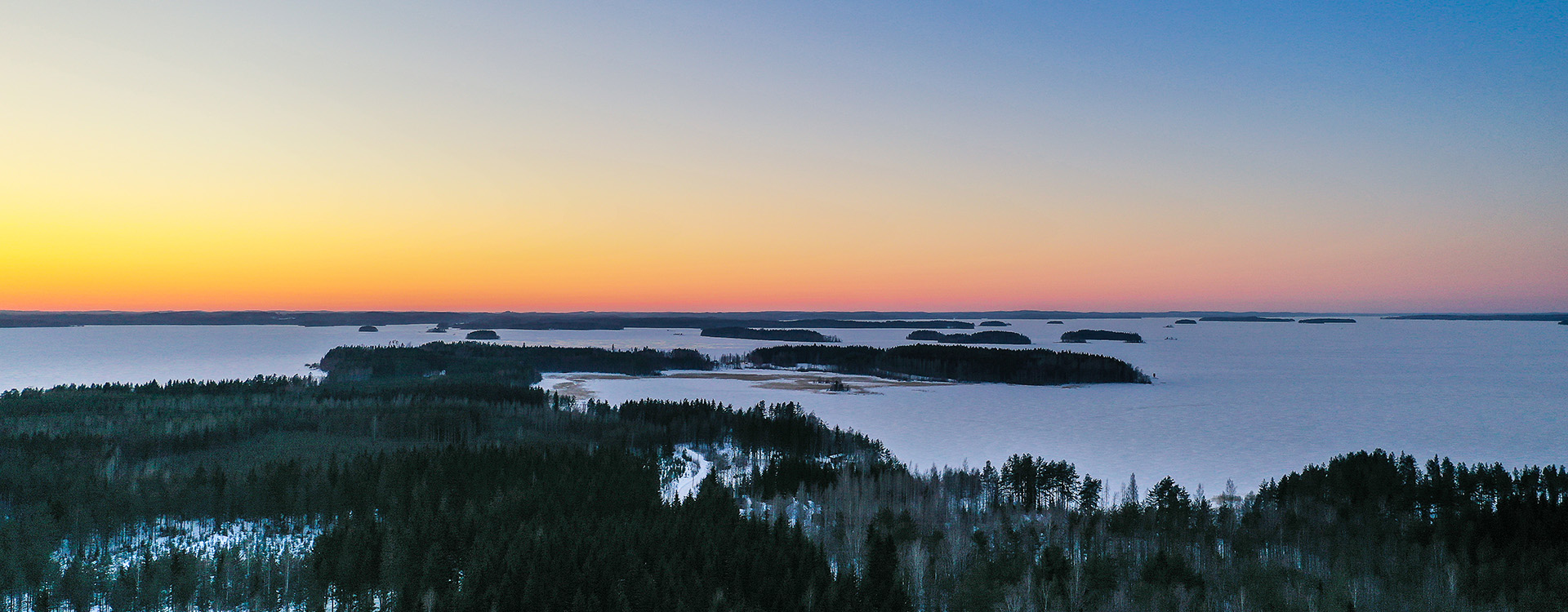 Talvinen järvimaisema dronella kuvattuna, auringon lasku taustalla, punertava taivaanranta, saaria ja vesi jäässä