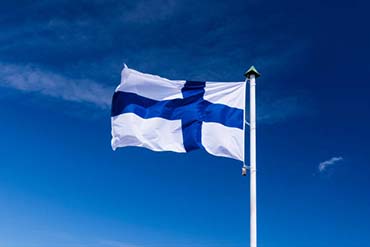 Kuvassa Suomen lippu. Lippu kuvattu ulkona ja taustalla näkyy sinistä taivasta.