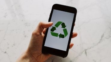 Ihmisen kädessä älypuhelin, jonka näytöllä vihreä kierrätys-merkki.